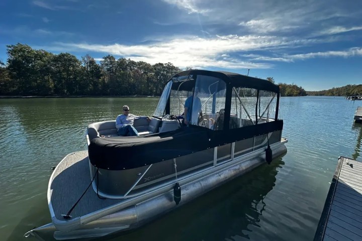 Pontoon boat with canopy on a lake, two people onboard, surrounded by trees, under a blue sky.