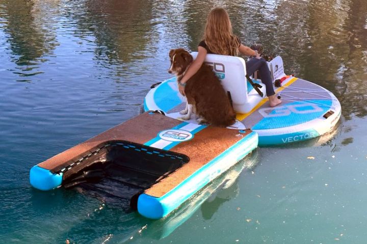 Person and dog sit on a floating platform on calm water.