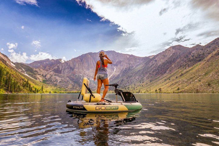 Person standing on a small boat in a lake surrounded by mountains under a partly cloudy sky.